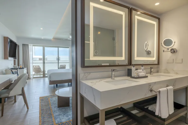 Double sinks and illuminated mirrors in a Luxury Junior Suite, with the bedroom and balcony beyond.