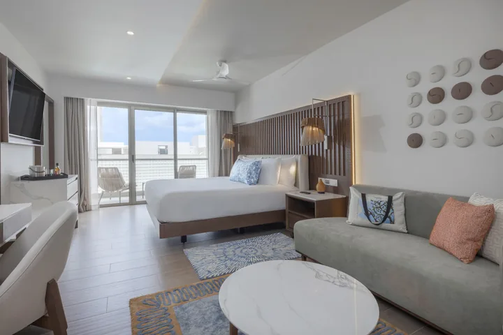 A king bed, sofa, round table and desk in a Royalton Vessence Barbados Luxury Junior Suite with balcony doors.