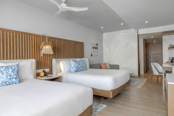 Two queen beds with white linens and blue pillows in a Royalton Vessence Barbados Luxury Junior Suite, with a sofa and desk.