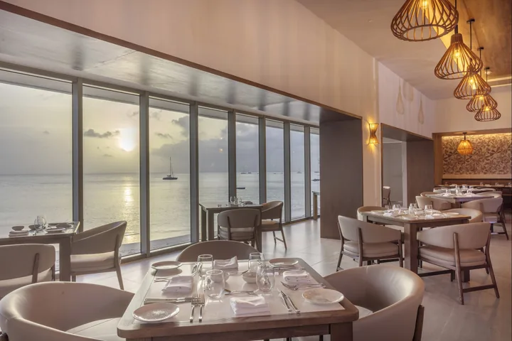 Set dining tables beneath pendant lights at La Scala, beside floor-to-ceiling windows overlooking the sea at sunset.