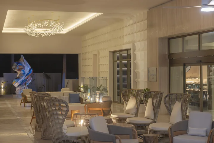 Woven chairs and sofas in the Royalton Vessence Barbados lobby seating area, with a chandelier and colorful sculpture.