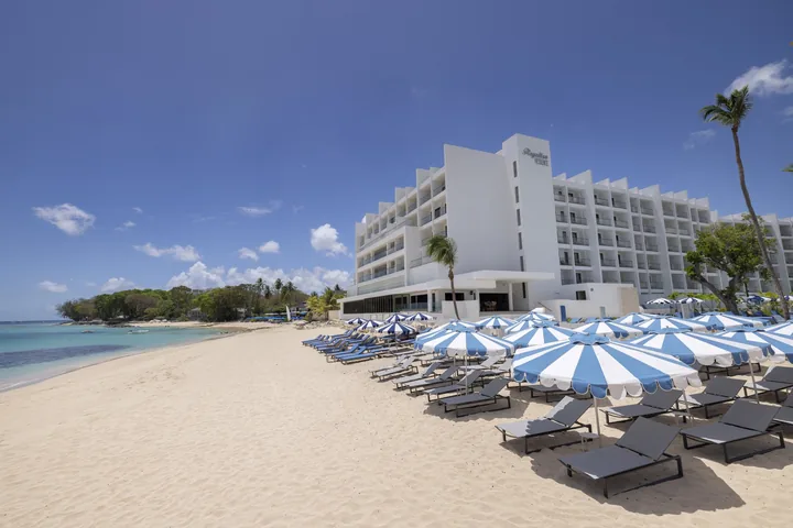 Sandy beach with loungers and blue-and-white umbrellas beside the Royalton Vessence Barbados resort and turquoise sea.